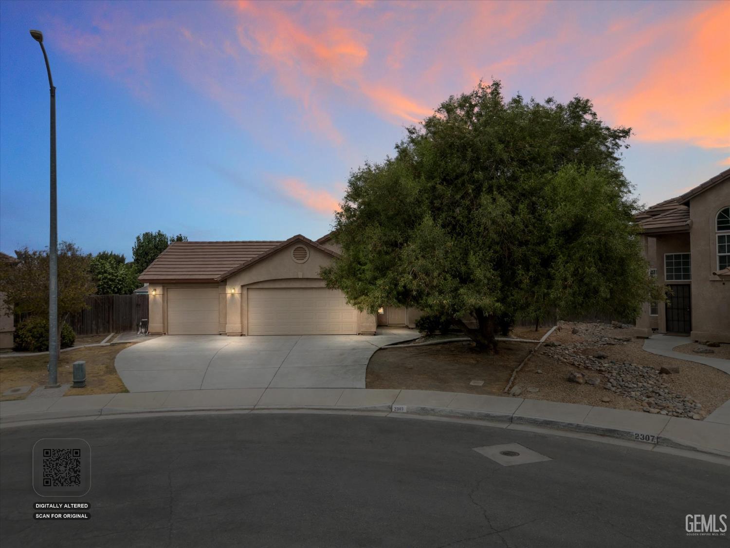 Beige stucco house with a two-car garage and large tree beneath a pink-and-blue sunset sky.