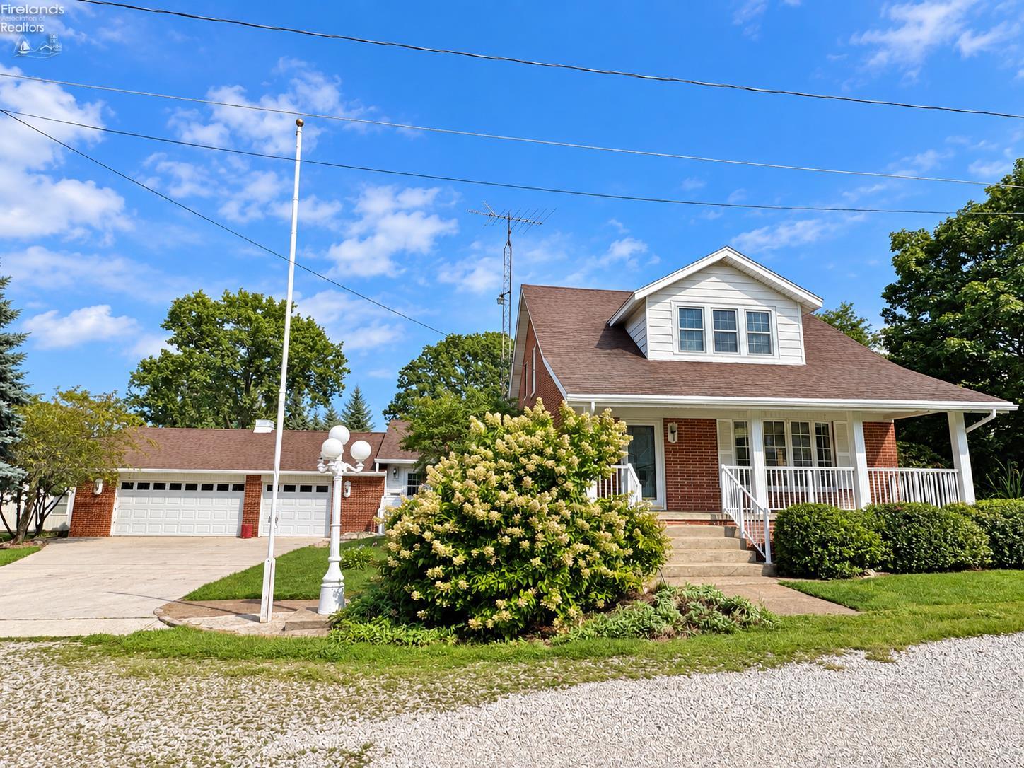 Brick house with a white dormer and wraparound porch, attached garage, and landscaped front yard.