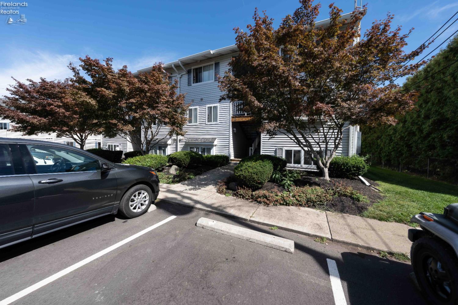 Gray-sided residential building behind autumn-colored trees, with a landscaped entrance and parking lot.